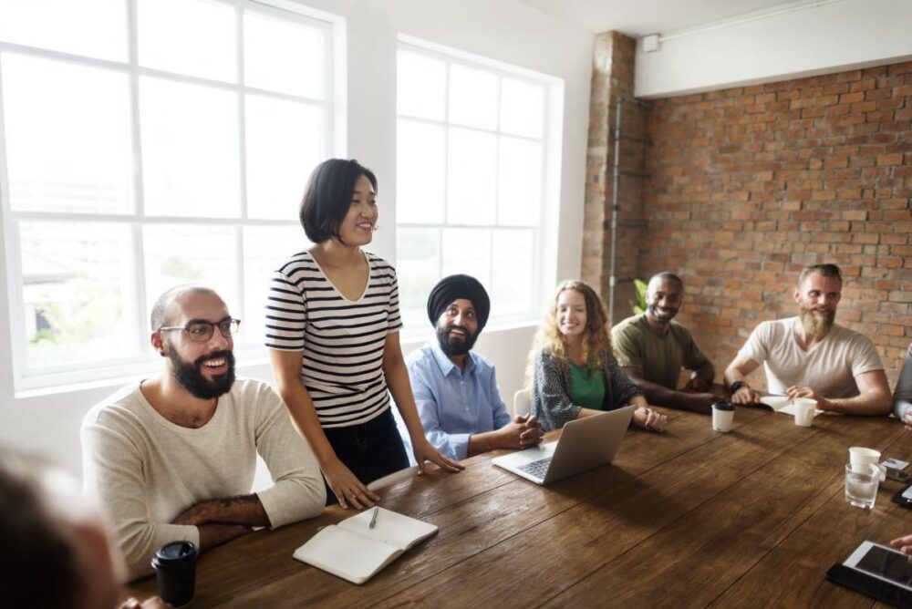 /group-of-colleagues-sitting-around-table-with-one-woman-standing-up-to-speak.jpg